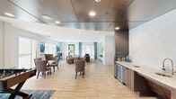 Located at 12639 Coit Rd in Dallas, TX, this single-floor community amenity space offers a thoughtfully designed environment that blends social, recreational, and functional areas seamlessly. The open-concept layout is anchored by light wood-look tile flooring that extends throughout, creating a warm and cohesive aesthetic. A striking curved dark metal ceiling feature defines the central lounge and game zone, contrasting beautifully with the bright white walls and recessed lighting found throughout the rest of the space. The main lounge area is furnished with plush seating arrangements, a built-in white media wall with a mounted flat-screen TV, and large windows dressed in sheer roller shades that flood the room with natural light and provide views to the outdoors. A dedicated game area includes a foosball table and casual seating clusters with tufted accent chairs around round tables. A sleek wet bar with a built-in beverage refrigerator and dark wood cabinetry serves as an entertainment hub. A private conference room features a solid wood dining-style table, upholstered chairs, a wall-mounted display, elegant pendant lighting, and a large window overlooking the surrounding greenery. The overall ambiance is modern yet welcoming, balancing upscale finishes with comfortable, functional design.