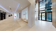 This single-floor lobby presents a striking contemporary design defined by soaring double-height ceilings and an abundance of natural light streaming through expansive floor-to-ceiling windows. The elongated corridor is lined with built-in seating alcoves recessed into crisp white walls, each fitted with cushioned benches, cylindrical bolster pillows, and articulating black wall sconces, offering intimate resting spots along the thoroughfare. Light-toned large-format tile flooring laid in a herringbone pattern runs throughout, amplifying the sense of openness.

A warm wood-slat ceiling with integrated linear lighting adds texture and warmth, contrasting beautifully with the otherwise all-white palette. One lounge area features a bold navy sectional sofa, a marble-topped coffee table, and sheer floor-to-ceiling drapery framing outdoor views of palm trees and the surrounding cityscape. A nearby dining area showcases a dramatic full-height marble accent wall, a natural wood communal table, and mixed modern seating. Curated artwork is displayed throughout the corridors. The elevator bank, finished in brushed stainless steel with dark surround panels and ambient blue accent lighting, anchors the space with a polished, modern presence.