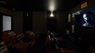 Located at 2001 Dogwood Dr in Fredericksburg, Virginia, this dedicated home theater room delivers an immersive, cinema-quality experience within a single-floor layout. The room is designed with a deliberately dark, dramatic aesthetic — deep charcoal and near-black walls absorb light and enhance contrast, creating an atmosphere reminiscent of a private screening room. Ambient wall sconces provide subtle, controlled illumination that sets the mood without disrupting the viewing experience. The seating arrangement consists of multiple rows of rich brown leather recliners, accented with striped throw pillows, and tiered to ensure unobstructed sightlines from every seat. A large flat-panel display serves as the focal point of the front wall, flanked by wall-mounted surround sound speakers strategically positioned throughout the room for balanced audio. Decorative macramé wall hangings and textured art panels add a personalized, bohemian touch to the otherwise sleek, dark interior. Recessed ceiling speakers further contribute to the multi-dimensional audio setup. A frosted-glass door provides a clean, private entrance to the space. The overall design is thoughtfully optimized for entertainment, combining comfort, acoustics, and visual style into a cohesive and functional private theater experience.