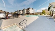 This outdoor Pool & Spa area is a well-appointed recreational amenity designed for relaxation and leisure. The centerpiece of the space is a generously sized swimming pool with clear, blue water, surrounded by a broad concrete deck that provides ample room for movement and lounging. White lounge chairs are positioned along the poolside, offering comfortable spots to unwind and enjoy the open sky above.

Adjacent to the pool is a dedicated hot tub spa, finished with tiled surrounds and equipped with stainless steel handrails for safe entry and exit. The bubbling waters of the spa provide a soothing retreat, and posted spa rules signage ensures safe usage for all guests.

The deck area is bordered by a building featuring a classic combination of brick wainscoting and horizontal lap siding in a neutral taupe tone, lending the space a clean, residential aesthetic. Sliding glass doors and windows along the building facade connect the outdoor deck to interior facilities, with convenient access points for users. Outdoor lighting fixtures are mounted on the building exterior to illuminate the space during evening hours.

The entire pool and spa area is enclosed by a black iron security fence with a gated entry, ensuring a safe and private environment. Trash receptacles and utility elements are thoughtfully integrated into the space. The surrounding residential buildings are visible beyond the fence, situating this amenity within a larger community setting.