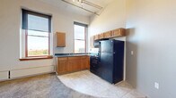 Located at 180 N Water St in downtown Rochester, NY, this one-bedroom, one-bathroom unit offers an efficient and functional layout across a single floor. The open-plan living, dining, and kitchen area serves as the heart of the space, characterized by notably tall ceilings and a rail lighting system that adds an industrial touch. The kitchen is well-appointed with warm-toned wood cabinetry, dark countertops, tile flooring, and black appliances including a refrigerator, range, microwave, and a double sink positioned beneath large windows that invite ample natural light and city views. The living and dining area flows seamlessly from the kitchen, featuring plush carpeting that adds warmth and comfort. The bedroom is a cozy, carpeted retreat with a large window offering additional urban views and a baseboard heating unit. Two closets provide convenient storage throughout the unit. A hallway connects the main living areas to the bathroom, which includes a full tub-shower combination, a vanity with matching wood cabinetry, and tile flooring consistent with the kitchen. The unit's elevated position provides notable city views, making it a well-situated residence in the heart of downtown Rochester.