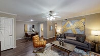 This single-floor residence offers a thoughtfully arranged layout connecting several distinct living areas with a cohesive, contemporary aesthetic. The living room serves as the central gathering space, featuring dark hardwood-style flooring, crown molding, and a ceiling fan with light fixture. A large statement artwork in gold and grey anchors one wall, complementing the warm neutral color palette. A wide window allows natural light to fill the room and provides an outdoor view.

Adjacent to the living area, the dining space accommodates a round table and seating, accented by matching artwork and crown molding details. The galley-style kitchen is outfitted with white shaker cabinetry, granite-style countertops, and stainless steel appliances, including a refrigerator, dishwasher, and range.

The bedroom offers plush carpeting, a ceiling fan, and a window with blinds that allows natural light. A secondary room functions as a flexible home office or study, featuring carpet, a ceiling fan, and a window. The bathroom includes a white vanity with granite-style countertops, a tub-shower combination, and wood-style flooring, completing this well-finished residence.