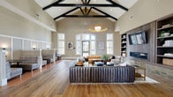 This expansive single-floor space is thoughtfully designed with a blend of modern and rustic architectural elements. The centerpiece is a grand living room showcasing soaring vaulted ceilings with striking exposed dark wood beam trusses, wide-plank hardwood flooring, and an abundance of natural light streaming through large arched and rectangular windows. A statement pendant light fixture anchors the seating area, which features plush sectional sofas and a built-in shelving unit flanking a fireplace with a mounted display screen. The dining room offers a refined gathering area highlighted by warm wood ceiling beams, an elegant chandelier, a shuffleboard table, and a built-in wet bar accented with green tile and pendant lighting. Comfortable booth-style seating with wainscoting walls provides an additional dining nook with warm sconce lighting. The hallway is clean and airy, finished with light hardwood flooring, framed artwork, and flush-mount lighting leading to exterior access points. The two bedrooms include a dedicated workspace nook with a marble-topped desk surface, a bold accent wall, and plaid carpeting. Throughout, a warm neutral color palette, curated art pieces, and high-end finishes create a polished, welcoming ambiance suited for both relaxation and social engagement.