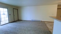 This single-floor space presents a straightforward, functional layout centered around an open main room. The room is finished with wall-to-wall carpet in a neutral gray-beige tone, providing a practical and comfortable underfoot surface. The walls are painted in a clean, bright white, contributing to a simple and airy atmosphere throughout the space. A baseboard heater runs along the lower portion of the far wall, offering an efficient heating solution integrated directly into the room's design. The entry door, finished in white with dark wood trim detailing along the door frame and floor baseboard, adds a subtle contrast to the otherwise uniform color palette. Multiple electrical outlets are positioned along the walls, offering practical utility. The ceiling is smooth and white, maintaining the overall clean aesthetic of the space. The open, uncluttered layout allows for flexible furniture arrangement, making it adaptable to a variety of uses. Overall, the space conveys a clean, minimalist character that serves as a blank canvas for customization.