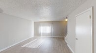 Located at 4322 Kostoryz Rd in Corpus Christi, Texas, this two-story residence offers a clean, neutral aesthetic throughout. The main living area greets visitors with light wood-style flooring, soft neutral-toned walls, and a textured popcorn ceiling. A large vertical-blind-covered sliding door floods the space with natural light, casting warm stripes across the floor. A staircase connects the two levels, adding a sense of vertical dimension to the layout. The kitchen is efficiently designed in a galley style, featuring white cabinetry, speckled laminate countertops, and a black electric range. A hallway from the kitchen leads directly to a bathroom, enhancing convenience and flow. The bedrooms are carpeted and well-lit, with one room offering a ceiling fan with integrated lighting, while another features a window with horizontal blinds that invites natural light. Additional living or flex space on the main floor also features the same wood-style flooring, open sightlines, and a pass-through view into the kitchen area. Throughout the home, consistent finishes including white trim, neutral paint, and tile-style flooring in wet areas create a cohesive and move-in-ready environment.