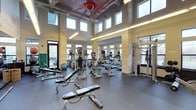 This fitness center is a thoughtfully designed workout facility that combines functionality with an open, energizing atmosphere. The space boasts soaring double-height ceilings with exposed industrial ductwork and a striking red-accented ceiling detail, lending the room a modern, urban character. Natural stone columns anchor the interior, adding a sense of solidity and architectural warmth that contrasts beautifully with the contemporary finishes throughout.

An abundance of natural light pours in through multiple rows of large windows positioned at both standard and clerestory heights, creating a bright and motivating environment throughout the day. The durable rubber flooring provides both safety and comfort underfoot, making it well-suited for high-activity use.

The facility is comprehensively outfitted with a wide array of cardio equipment, including treadmills, elliptical machines, and stationary bikes — many featuring integrated display screens. Strength training is well represented with adjustable weight benches, free weights, and a multi-station cable machine, offering a complete workout experience. The spacious layout ensures ample room between equipment, allowing for comfortable and unrestricted movement. This fitness center is designed to meet the needs of a broad range of users, from casual exercisers to dedicated athletes.