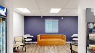 This single-floor space is thoughtfully designed to serve as a comfortable and functional communal hub. The layout flows seamlessly between distinct zones, each with its own purpose and character. The lounge area makes an immediate impression with a bold navy blue accent wall as its backdrop, warm wood-look flooring, and a striking amber velvet sofa flanked by white bouclé accent chairs with cane detailing — creating an inviting and stylish place to relax or socialize. Natural light filters through horizontal-blind windows, complemented by recessed panel ceiling lighting throughout.

Adjacent to the lounge, a retail-style snack and beverage section features fully stocked display shelving and a glass-door Pepsi refrigerator, offering convenient access to drinks and packaged goods. A cozy game nook provides a quieter retreat, furnished with cane-back wooden chairs and a charming chess table, ideal for casual leisure.

The kitchen area is well-appointed with light wood cabinetry, granite-look countertops, a stainless steel range, dishwasher, and full-size refrigerator, along with a sink and ample storage. A bistro-style high-top table with gold-accented stools adds a casual dining option. The consistent wood-look flooring and bright, even lighting throughout the space tie all areas together into a cohesive, welcoming environment.