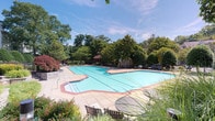 This outdoor amenity space centers around a generously sized swimming pool with a distinctive freeform shape, edged with a classic red brick border that adds a touch of traditional character. The pool deck is finished in light-toned concrete, providing ample space for lounge chairs and seating arrangements that invite relaxation. A covered gazebo or pavilion structure is visible in the background, offering a shaded retreat for poolside gatherings. The surrounding landscape is rich with mature deciduous trees and well-maintained greenery, creating a naturally enclosed and private atmosphere. Lush plantings frame the perimeter of the pool area, enhancing the sense of seclusion while adding vibrant color and texture throughout the seasons. Seating areas, including tables and chairs, are positioned around the deck to accommodate social gatherings. The clear blue water of the pool reflects the open sky above, contributing to the bright and inviting ambiance of the space. Overall, the area presents a well-appointed outdoor recreational environment that balances functional amenities with a naturally serene and park-like setting.