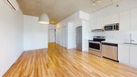 Located at 205 Rue De L'Équinoxe in Brossard, QC, this modern unit offers a thoughtfully designed single-floor layout that maximizes both function and style. The open-concept living area is defined by warm hardwood flooring throughout and striking exposed concrete ceilings, lending an industrial-chic character to the space. Large floor-to-ceiling windows and a sliding glass door flood the interior with abundant natural light while opening onto a private balcony with expansive city views of the surrounding Brossard skyline.

The kitchen is seamlessly integrated into the living area and is outfitted with sleek white cabinetry, a stainless steel refrigerator, electric range, and built-in microwave — a clean, functional setup well-suited for everyday use. A pendant light fixture anchors the open living and dining zone, adding a contemporary design touch. Sliding doors along one wall provide access to in-unit laundry, adding practical convenience.

The bathroom is finished in an all-white palette, featuring a full bathtub with tiled surround, a modern floating vanity, and a well-placed mirror. The cohesive, minimalist design is carried throughout every room, creating a bright and airy atmosphere. A hallway with a closet connects the living space to the bathroom, completing this efficiently planned home.