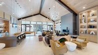 Located at 100 Dorchester Dr in Lumberton, NJ, this thoughtfully designed single-floor community space offers a generously proportioned, open-concept layout that seamlessly blends modern aesthetics with functional comfort. The most striking architectural feature is the dramatic vaulted ceiling with exposed dark wood beams and a light wood paneled surface, which lends the space a warm yet contemporary character. Two statement chandeliers — one a sculptural, branch-like fixture and another featuring cascading globe pendants — serve as focal points throughout the open floor plan. Large floor-to-ceiling windows along one wall flood the interior with abundant natural light while providing views to the exterior. The main lounge area is anchored by multiple seating arrangements, including plush sofas and accent chairs, centered around a sleek fireplace set against a bold blue-gray feature wall with built-in shelving. An island-style kitchenette with bar seating provides a social gathering hub. The entry corridor showcases a dramatic textured accent wall in a geometric pattern, illuminated by globe sconces, alongside a built-in bench and curated display shelving. A billiards table and a mirrored hallway add to the variety of amenities. Large-format tile flooring flows throughout, tying all areas together cohesively.