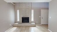 This two-story home offers a well-defined layout spread across two evenly sized floors. The main level opens into a combined living, dining, and kitchen area centered around a striking floor-to-ceiling whitewashed brick fireplace flanked by narrow vertical windows that draw in natural light. Light ceramic tile flooring flows throughout the main level, complementing the neutral gray walls. A set of French doors provides additional brightness and access to the exterior. The kitchen features dark espresso cabinetry, a double-basin sink, a tile countertop with a breakfast bar extension, a white electric range, and track lighting overhead. A half bathroom on the main floor adds convenience for guests, finished with tile flooring and a vanity with a stone-toned countertop. A staircase with wrought iron railings leads to the upper level, where plush carpet extends throughout. The generously sized primary bedroom includes a ceiling fan, a large window with vertical blinds, and a dedicated closet. The full bathroom on this floor is well-appointed with large-format tile surround, a soaking tub, a decorative mosaic accent border, and a stone-toned vanity countertop. The upper landing offers an open railing overlooking the fireplace wall below, adding architectural depth to the home.