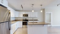 Located at 11901 S Freedom Park Dr in Herriman, Utah, this single-floor residence offers a clean, modern design with a neutral color palette throughout. The open-concept kitchen serves as the heart of the home, showcasing crisp white cabinetry, dark quartz-style countertops, and a full suite of stainless steel appliances including a refrigerator, electric range, over-the-range microwave, and dishwasher. A central island with an integrated sink provides additional workspace and casual seating, illuminated by stylish pendant lighting and complemented by track lighting overhead. Light wood-style flooring runs throughout the main living and kitchen areas, creating a cohesive and airy feel. The living area benefits from natural light through large windows fitted with blinds. Bedrooms feature plush carpeting, ceiling fans, and windows that invite natural light. The bathroom is finished with light tile flooring, white vanity cabinetry with a dark countertop, and a full tub and shower combination. A dedicated laundry room provides added convenience with side-by-side washer and dryer units and built-in shelving above. The overall ambiance is bright, functional, and move-in ready, with consistent modern finishes throughout.