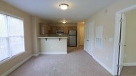 This single-floor residence offers a practical and welcoming open-concept layout that seamlessly connects the living area and kitchen. The living space is finished with plush, neutral-toned carpeting that adds warmth and comfort underfoot, while the clean white baseboards and soft beige walls create a bright, cohesive aesthetic throughout.

The kitchen is a functional centerpiece, anchored by a peninsula counter with an overhanging bar that provides casual seating and defines the space without interrupting the open flow. It is equipped with stainless steel appliances, including a full-size refrigerator, a microwave, and a range, all complemented by medium-toned wood cabinetry that offers generous storage. Flush ceiling fixtures provide consistent overhead lighting across both the kitchen and living zones.

A white paneled door with a closet-style bi-fold configuration is visible along one wall, offering practical storage access directly from the main living area. A hallway leads toward additional rooms, suggesting a thoughtful floor plan with clear separation between living and private spaces. The overall ambiance is clean, understated, and move-in ready, making it a highly functional space suited for comfortable everyday living.