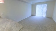 This single-floor space offers a clean, minimalist aesthetic defined by bright white walls and light-toned carpeted flooring throughout. The interior is bathed in natural light, courtesy of a large window dressed with vertical blinds that allow for flexible light control while maintaining a sense of openness. The architectural design is straightforward and functional, with smooth wall finishes and crisp white baseboards that contribute to a fresh, well-maintained appearance. The open layout provides a versatile living area with ample room to accommodate a variety of furniture arrangements. A partial view of what appears to be a stone or granite surface in the foreground suggests the presence of a countertop or decorative element, adding a touch of texture and contrast to the otherwise neutral palette. The overall ambiance of the space is calm and airy, making it well-suited for those who appreciate a clean, uncluttered environment. The combination of natural light, neutral tones, and open floor space creates an inviting and adaptable interior ready to be personalized.
