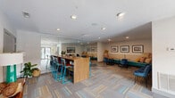 Located at 936 Mountain Creek Rd in Chattanooga, Tennessee, this community lounge is a thoughtfully designed gathering space that offers a welcoming and functional environment for socializing, working, and relaxing. The single-floor layout is organized into distinct zones, each serving a different purpose while maintaining a cohesive, contemporary aesthetic throughout.

The space features multi-toned carpet tile flooring in warm earth and cool blue tones, adding visual texture and defining the various seating areas. A full-length floor-to-ceiling mirror wall runs along one side, amplifying the sense of openness and reflecting natural light that filters in through large glass doors leading to an outdoor pool area.

A built-in banquette lined with plush cushions and colorful accent pillows provides a cozy lounge nook, complemented by round wood-topped tables and teal velvet chairs. Nearby, a pair of cushioned armchairs and additional round tables create a more casual conversation area. A large communal high-top table with striking teal-framed bar stools serves as a collaborative workspace or social hub. Recessed lighting illuminates the clean white ceilings throughout.

Framed artwork depicting local Chattanooga scenes adds a regional character to the walls. Near the entry, a coffee kiosk and beverage vending machines offer added convenience. The overall ambiance is modern, colorful, and inviting.