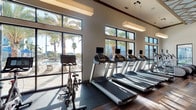 This single-floor fitness center is a thoughtfully designed workout facility that combines contemporary architecture with a comprehensive suite of exercise equipment. The space is divided into two distinct zones: a bright, window-lined cardio area and a dedicated strength training section, each offering a distinct workout environment.

The cardio zone is bathed in natural light streaming through large floor-to-ceiling windows and clerestory windows above, offering views of swaying palm trees and an outdoor pool area. A row of treadmills with individual screen consoles and stationary cycling bikes are arranged along the windowed wall, creating an energizing atmosphere for aerobic exercise. Rich hardwood-style flooring runs throughout, complemented by rubber fitness flooring beneath the equipment.

The strength training area features an impressive geometric latticed ceiling detail, lending architectural interest to the space. Wood-paneled accent walls add warmth and texture, while full-length mirrors amplify the sense of space. This zone is equipped with a cable machine, weight benches, a multi-station strength machine, and a dumbbell rack. Pendant lighting fixtures provide focused illumination throughout.

Access points include glass entry doors that connect to the surrounding outdoor community. The overall ambiance is bright, open, and modern — well-suited for a full range of fitness activities.
