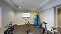 Located at 5721 Goodstone Dr in Raleigh, NC, this single-floor fitness center offers a well-appointed facility designed to support a comprehensive workout experience. The main exercise area features a wide selection of strength-training machines, free weights, and resistance equipment arranged across a durable, rubber-composite flooring surface. Recessed lighting and ceiling fans ensure the space remains bright and well-ventilated, while large wall-mounted mirrors expand the sense of space and allow users to monitor their form during workouts. A wall-mounted television adds convenience for entertainment during training sessions. An adjacent studio room provides a dedicated space for cardio and group fitness activities, equipped with stationary cycling bikes, stability balls, resistance bands, yoga mats, and medicine balls neatly organized on storage racks. This room also features a large wall-mounted display screen and a tray ceiling with recessed lighting, creating a focused and motivating environment for guided workouts. Together, the two areas form a cohesive and functional fitness facility suited for a wide range of exercise routines and fitness levels.