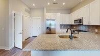 Located at 1841 Dusty Miller Ln in Wilmington, NC, this single-floor residence offers a well-designed layout with modern finishes and a clean, neutral aesthetic throughout. The kitchen is a standout feature, showcasing tall white shaker cabinetry, a tasteful tile backsplash, granite countertops, and a full suite of stainless steel appliances including a French door refrigerator, range, and built-in microwave. A large island with an undermount sink and matte black faucet provides additional prep space and a natural gathering point. Wood-look flooring runs through the main living and kitchen areas, adding warmth and continuity. The living area is generously sized with soft carpet underfoot and a ceiling fan overhead, flowing seamlessly from the kitchen. Bedrooms are carpeted and feature neutral walls, ceiling fans, and ample natural light from well-placed windows. The primary bedroom connects to a walk-in closet outfitted with multi-tier wire shelving for versatile storage. The bathroom is finished with white subway tile surrounding a full soaking tub and shower combination, complemented by granite countertops, wood-look flooring, and matte black fixtures. The overall ambiance is bright, clean, and move-in ready.