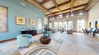 This single-floor community clubhouse is defined by its sophisticated architectural design and thoughtfully appointed spaces. The centerpiece is a grand lounge area with soaring coffered ceilings accented by rich wood beams, recessed lighting, and transom windows that invite abundant natural light. Stone tile flooring, decorative area rugs, and teal accent walls create a cohesive, welcoming palette throughout. Comfortable seating arrangements, curated artwork, and elegant furnishings give this space a refined yet approachable ambiance.

The reception entry features glass-paneled double doors, a welcoming desk, and tall windows that frame views of the surrounding outdoor landscape. Adjacent areas include a casual dining nook with a wall-mounted TV and natural light from wood-trimmed windows, and a dedicated conference room outfitted with a large boardroom table, executive seating, and an arched window overlooking the exterior grounds.

A curved wet bar serves as a social hub, complete with a granite countertop, pendant lighting, built-in shelving, stainless steel appliances, and a mosaic tile backsplash. Throughout the facility, consistent use of stone tile flooring, warm wood trim, and teal accent walls ties each area together into a polished and functional whole.