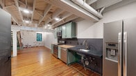 Located at 101 E Walnut St in downtown Rogers, AR, this two-story loft blends industrial character with modern updates. The main floor serves as the primary living and kitchen area, showcasing exposed white-painted wood beam ceilings with visible ductwork, warm hardwood floors, and a striking exposed brick accent wall. The kitchen is outfitted with sage green cabinetry, dark tile backsplash, stainless steel appliances, and a butcher block countertop insert, offering both style and functionality. A staircase with sage green treads, wood balusters, and cable railing connects the two floors, adding a contemporary design element. The upper level houses the spacious primary bedroom, featuring exposed beam ceilings, a ceiling fan, shiplap accent wall, and a sliding barn door leading to a walk-in closet with dual hanging rods and shelving. The bathroom is a standout, finished with white subway tile walls, hexagonal floor tile, a glass-enclosed walk-in shower, a vessel sink atop a wood and metal vanity, and matte black fixtures. A utility room completes the layout. Throughout the space, the interplay of raw materials and refined finishes creates a cohesive, urban-industrial ambiance ideal for modern living.