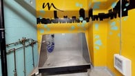The Tanner Flats Dog Wash is a purpose-built, single-level self-service pet grooming facility designed with both functionality and personality in mind. The space is immediately eye-catching, with bold yellow walls adorned with playful teal bubble graphics and a striking black city skyline mural that wraps around the room. Paw print decals and a prominent "#dogwash" graphic reinforce the space's fun, pet-forward identity.

At the heart of the facility are deep, commercial-grade stainless steel wash tubs equipped with coiled blue hoses and mounted fixtures, providing a practical and easy-to-clean setup for bathing pets of all sizes. Alongside the tubs, a grooming table with a non-slip surface offers a dedicated area for drying and finishing. A wall-mounted paper towel and supplies dispenser keeps essentials within easy reach.

The structural walls are constructed of painted concrete block in a complementary mint green, giving the space a clean, utilitarian foundation. Exposed copper plumbing lines run along the walls, adding an industrial character to the design. A wall-mounted ventilation fan ensures proper airflow, while a posted rules and instructions sign keeps users informed. A dedicated dog-access entry door provides convenient, controlled access to the facility. The overall ambiance is energetic, clean, and welcoming for pet owners.