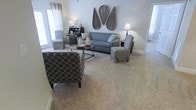 This single-floor living space presents a welcoming and cohesive design centered around a neutral color palette of grays, taupes, and whites. The spacious living area is anchored by plush, light-toned wall-to-wall carpeting that adds warmth underfoot. Large windows dressed with white curtains allow generous natural light to fill the room, enhancing its airy and open feel. The seating arrangement features a gray upholstered sofa accented with teal throw pillows, complemented by a patterned accent chair in a bold circular motif and slipcovered side chairs in soft gray tones. A round wrought-iron coffee table serves as a functional centerpiece, styled with decorative vases. Wall décor includes a distinctive pair of woven rattan leaf-shaped pieces that add a natural, organic texture to the space. A dark wood console table with a lamp and framed accents anchors one corner, contributing depth and contrast. The overall ambiance is calm, coordinated, and comfortable, reflecting a thoughtfully styled interior suited for relaxed everyday living.