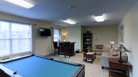 This single-floor amenity lounge is designed to offer a well-rounded recreational and social environment. The centerpiece of the room is a full-sized pool table with a blue felt surface, positioned prominently to anchor the space. Flush-mounted fluorescent light panels illuminate the room evenly, complementing the natural light that filters in through the windows dressed with white curtains.

Along one wall, a wall-mounted flat-screen TV sits at a comfortable viewing height, while nearby, a dark wood bar-height desk with two matching stools provides a casual workspace or social spot. A tall dark wood bookshelf, stocked with books and small decorative items, adds a touch of warmth and character to the corner.

On the opposite side of the room, a cozy seating area features a cushioned armchair and a sofa arranged around a wooden coffee table, creating an inviting lounge nook. Light neutral tones on the walls and carpet contribute to a relaxed, welcoming atmosphere throughout. The layout is open and functional, making the space suitable for both active recreation and quiet leisure.