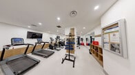 Located at Halifax St in Burnaby, BC, this fitness facility offers a well-appointed exercise room designed to support a full range of workout routines. The space features a clean, modern aesthetic with bright white walls, recessed pot lighting, and a light-toned resilient floor that creates an open, energizing atmosphere.

The cardio zone is prominently equipped with multiple treadmills, elliptical trainers, and a stair climber, each positioned along a wood-accented accent wall with wall-mounted flat-screen televisions for entertainment during workouts. A rowing machine and stationary cycle round out the cardio options.

The strength training area includes a multi-functional cable machine system, a dedicated free weight section with a full rack of dumbbells neatly organized along a floor-to-ceiling mirror wall, a weight bench, and a leg press/extension machine. Kettlebells and medicine balls are also available for functional training, alongside stability balls and resistance accessories.

Full-length mirrors are strategically placed to support proper form and enhance the sense of space throughout the room. The layout is thoughtfully organized to allow multiple users to exercise simultaneously without crowding, making this a practical and comprehensive amenity for residents of the building.