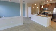 This single-floor space features an open-concept layout that connects the living area to the kitchen, creating a sense of spaciousness and ease of movement throughout. The living area is finished with plush neutral carpeting and is highlighted by a soft blue accent wall, adding a calming visual contrast to the warm beige tones that dominate the rest of the space. Structural columns with clean white trim define the transition between zones without fully separating them, maintaining an airy, connected feel.

The kitchen is well-appointed with rich, warm-toned cabinetry and granite countertops that extend into a peninsula or breakfast bar, offering both additional prep space and casual seating potential. Pendant lighting hangs above the counter, providing focused task lighting while contributing to the overall aesthetic. Additional track lighting on the ceiling ensures the kitchen remains well-lit. Stainless steel appliances are visible in the background, complementing the cabinetry. A white paneled door leads to an adjoining room, suggesting further functional spaces beyond what is immediately visible. Overall, the space conveys a clean, modern, and comfortable ambiance.