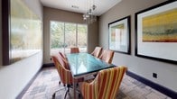 This single-floor meeting space is thoughtfully designed to support professional gatherings in a polished yet welcoming environment. The central feature is a rectangular glass-top conference table surrounded by upholstered chairs in warm striped fabric — a combination of red, gold, and green tones — that adds a layer of comfort to the functional setting. A cluster of modern pendant lights hangs above the table, complementing the recessed lighting throughout the space.

Natural light enters generously through large windows overlooking a landscaped outdoor area, creating a bright and pleasant atmosphere. Abstract artwork in warm earth tones is displayed on the neutral-toned walls, contributing to a refined aesthetic. The patterned carpet, featuring geometric motifs in muted hues, adds visual texture underfoot.

Adjacent to the main meeting area is a built-in service counter finished in rich dark wood cabinetry topped with dark granite. This area includes a printer and is separated from the corridor by a multi-pane interior window, offering visibility while maintaining distinct zones. The overall design balances functionality and style, making it a well-equipped and professional space for meetings or presentations.
