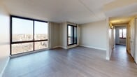 Situated along Memorial Drive in Cambridge, Massachusetts, this residence offers a thoughtfully designed single-floor layout with a clean, contemporary aesthetic throughout. The living area is a standout feature, boasting expansive floor-to-ceiling windows that frame sweeping elevated views of the Cambridge and Boston skyline, flooding the space with abundant natural light. Light-toned hardwood-style flooring runs throughout the main living areas, complementing the neutral, warm wall tones and textured tile ceilings that contribute to the space's modern feel. The open living and sleeping areas provide generous room for flexible furnishing arrangements, benefiting from the panoramic window walls on multiple sides. The kitchen is well-equipped with crisp white cabinetry, ample upper and lower storage, light countertops, a stainless steel sink, and black appliances including a range and dishwasher, offering both functionality and a clean visual contrast. The bathroom features a vanity with an integrated sink, a large mirror, and a combined tub and shower with decorative curtain. The overall ambiance of the unit is bright, open, and urban, making it an ideal space for those seeking a comfortable residence with commanding views in the heart of Cambridge.