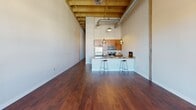 This single-floor loft-style unit showcases an industrial-chic design characterized by dramatically high ceilings with exposed wooden beams and visible ductwork, creating an open, airy atmosphere throughout. Rich, dark hardwood flooring extends across the main living area, reflecting natural light that streams in through multiple windows offering urban views.

The open-concept kitchen serves as a central feature, equipped with a full suite of stainless steel appliances, including a refrigerator, range, and built-in microwave. Warm-toned wood cabinetry lines the walls and reaches toward the ceiling, maximizing storage. Granite countertops continue across a peninsula breakfast bar, illuminated by pendant lighting and accommodating bar-stool seating—ideal for casual dining or entertaining.

The spacious living area benefits from soaring ceiling height and an open layout that allows for versatile furniture arrangements. A separate carpeted room provides a quieter, more private space suitable for use as a bedroom or home office, with an access panel visible on the wall.

The bathroom features a generous layout with a wood-toned vanity, granite countertop, single sink, large mirror, tile flooring, and a shower-tub combination with a curtain enclosure. Grab bars are installed, reflecting accessible design considerations throughout the space.