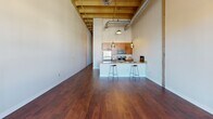 This single-floor loft-style unit showcases an industrial-chic design characterized by dramatically high ceilings with exposed wooden beams and visible ductwork, creating an open, airy atmosphere throughout. Rich, dark hardwood flooring extends across the main living area, reflecting natural light that streams in through multiple windows offering urban views.

The open-concept kitchen serves as a central feature, equipped with a full suite of stainless steel appliances, including a refrigerator, range, and built-in microwave. Warm-toned wood cabinetry lines the walls and reaches toward the ceiling, maximizing storage. Granite countertops continue across a peninsula breakfast bar, illuminated by pendant lighting and accommodating bar-stool seating—ideal for casual dining or entertaining.

The spacious living area benefits from soaring ceiling height and an open layout that allows for versatile furniture arrangements. A separate carpeted room provides a quieter, more private space suitable for use as a bedroom or home office, with an access panel visible on the wall.

The bathroom features a generous layout with a wood-toned vanity, granite countertop, single sink, large mirror, tile flooring, and a shower-tub combination with a curtain enclosure. Grab bars are installed, reflecting accessible design considerations throughout the space.