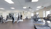 Located at 100 Millspring Dr in Durham, North Carolina, this fitness center offers a well-equipped, single-floor space designed to support a comprehensive workout experience. The interior features a drop-panel ceiling with recessed lighting panels and ceiling fans that ensure consistent air circulation and a comfortable exercise environment throughout. Large windows along the exterior wall invite natural light into the space, enhancing the open and airy atmosphere. The flooring is finished with durable rubber gym matting, providing both safety and functionality for a wide range of exercises. The strength training area is outfitted with a multi-station cable and weight machine, complemented by full-length wall mirrors that visually expand the space and allow users to monitor their form. The cardio section is lined with a row of modern elliptical machines and at least one stationary cycling bike, accommodating multiple users simultaneously. The neutral, light-toned walls and clean layout give the facility a tidy, professional appearance. Overall, the space is thoughtfully organized to balance cardio and strength training zones, creating a functional and welcoming environment suitable for residents or members looking for a convenient on-site fitness option.