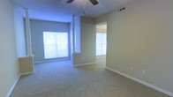 This single-floor residence offers a welcoming and functional layout designed for comfortable everyday living. The main living area is generously proportioned, featuring soft wall-to-wall carpeting underfoot and neutral, warm-toned walls that create a calm and inviting atmosphere. A ceiling fan is centrally mounted overhead, providing both practical airflow and a subtle design touch.

Large windows along the exterior walls flood the space with abundant natural light, offering views of the surrounding outdoor greenery and enhancing the open, airy feel of the interior. The windows are fitted with roller blinds, allowing for adjustable light control and privacy.

A partial column or architectural divider subtly defines the transition between the main living area and an adjoining room, which is accessible through an open doorway. This connected space mirrors the same bright, carpeted aesthetic, maintaining a cohesive design flow throughout the home. White baseboards provide a clean contrast against the neutral walls, adding a crisp, finished look to each room.

The overall ambiance of the space is light, understated, and move-in ready, with a straightforward layout that lends itself well to a variety of furnishing arrangements and personal styles. The single-floor design ensures easy accessibility and seamless movement throughout the home.