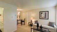 This single-story residence in Arlington, Texas, presents an open, connected layout that seamlessly integrates the living and dining areas into one cohesive space. The living area features plush wall-to-wall carpeting in a warm beige tone, contributing to a comfortable and inviting atmosphere. A glass-top coffee table anchors the seating area, complemented by neutral-toned furnishings and decorative accents that enhance the understated elegance of the room. A framed piece of wall art adds a finishing touch to the living space.

Adjacent to the living area, the dining space accommodates a compact bistro-style table with matching chairs, illuminated by a pendant light fixture that provides focused, warm lighting. The transition between these two areas is fluid, making the overall layout feel open and functional. The walls throughout are finished in a soft, neutral palette, reinforcing a calm and cohesive aesthetic.

A hallway visible from the main living space leads to additional areas of the home. The textured ceiling adds subtle architectural character. Overall, the space offers a practical and well-appointed single-floor layout suited for comfortable everyday living.