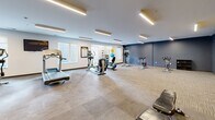 Located at 101 Main St in Lafayette, Indiana, this fitness center offers a generously sized, single-floor exercise room designed to support a well-rounded workout routine. The space features a clean, modern aesthetic with a neutral color palette accented by bold dark gray and navy blue walls, complemented by eye-catching yellow directional arrow graphics that add an energetic, motivational feel to the environment.

The exercise room is outfitted with a comprehensive selection of cardio equipment, including treadmills, elliptical machines, a recumbent bike, upright stationary bikes, and spinning cycles. A dedicated free weights area with a dumbbell rack and adjustable bench provides options for strength training, while a vertical knee raise tower supports bodyweight and core exercises. Colorful stability balls add versatility to the fitness offerings.

Large windows along the exterior walls allow natural light to filter into the space, offering views of the surrounding downtown Lafayette streetscape. Recessed linear LED lighting throughout ensures consistent, well-lit conditions at any time of day. The flooring is a durable, low-pile carpet tile suited for fitness use, and a wall-mounted television is positioned for easy viewing during workouts. A water station and posted fitness rules round out the practical amenities, making this a fully functional and inviting exercise facility.