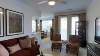 This single-floor residence showcases an open-concept layout that connects the living, dining, and kitchen areas in a fluid, functional arrangement. The living area is anchored by plush, patterned accent chairs with matching ottomans and a deep red sofa adorned with decorative pillows, creating a warm and inviting atmosphere. A ceiling fan with integrated lighting adds both comfort and practicality overhead, while crown molding lends a refined architectural touch to the space.

A half-wall partition gracefully separates the kitchen from the main living area, preserving an open feel while defining each zone. The kitchen features pendant lighting that adds a modern accent above the counter. The dining area sits just beyond, bathed in natural light from a large bay window dressed with blinds and flowing drape curtains.

French doors lead to an outdoor area, allowing generous natural light to pour into the space and offering a seamless indoor-outdoor connection. Neutral-toned carpet throughout the living and dining areas complements the warm color palette of the furnishings. Overall, the space balances comfort and style with thoughtful design details and an abundance of natural light.