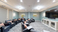 Located at 1470 S Havana St in Aurora, CO, this well-appointed screening and media lounge room offers a comfortable and inviting environment designed for group viewing and relaxation. The single-floor space is thoughtfully arranged with multiple dark leather recliner chairs, each equipped with built-in side tables and cup holders, and accented with warm peach-toned throw pillows for added comfort. A large flat-screen television is mounted on one wall, complemented by a white cabinet console beneath it that provides both display space and storage. The room's soft teal-blue walls create a calm, soothing atmosphere, while patterned gray carpeting adds a sophisticated texture underfoot. Overhead, a drop-tile ceiling is outfitted with a combination of flush-mount drum light fixtures and recessed lighting, ensuring the space is evenly and warmly illuminated. Wall sconces provide additional ambient lighting along the perimeter. Colorful landscape paintings adorn the walls, contributing an artistic and cheerful character to the room. Louvered doors on one wall offer a refined architectural detail. The overall design strikes a balance between comfort and style, making this space well-suited for community screenings, presentations, or casual group gatherings.