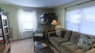 This single-floor space presents a welcoming living area designed with comfort and functionality in mind. The room features neutral-toned walls complemented by plush carpeted flooring, creating a warm and inviting atmosphere. Natural light fills the space through multiple windows dressed with white sheer curtains and horizontal blinds, offering both brightness and privacy.

The living area is thoughtfully arranged with a generously sized upholstered sofa in a muted taupe tone, paired with a coordinating patterned accent chair that adds visual interest to the space. A dark-finished rectangular coffee table anchors the seating arrangement, providing a practical surface for everyday use. A matching end table holds a classic table lamp, contributing to the room's warm ambient lighting.

A decorative round mirror serves as a focal wall accent, positioned above a tall potted plant that introduces a natural element to the interior. The overall design aesthetic is casual and understated, combining functional furnishings with simple decorative touches to create a comfortable, livable environment suited for relaxing or entertaining.