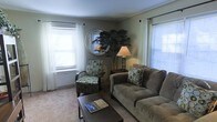 This single-floor space presents a welcoming living area designed with comfort and functionality in mind. The room features neutral-toned walls complemented by plush carpeted flooring, creating a warm and inviting atmosphere. Natural light fills the space through multiple windows dressed with white sheer curtains and horizontal blinds, offering both brightness and privacy.

The living area is thoughtfully arranged with a generously sized upholstered sofa in a muted taupe tone, paired with a coordinating patterned accent chair that adds visual interest to the space. A dark-finished rectangular coffee table anchors the seating arrangement, providing a practical surface for everyday use. A matching end table holds a classic table lamp, contributing to the room's warm ambient lighting.

A decorative round mirror serves as a focal wall accent, positioned above a tall potted plant that introduces a natural element to the interior. The overall design aesthetic is casual and understated, combining functional furnishings with simple decorative touches to create a comfortable, livable environment suited for relaxing or entertaining.