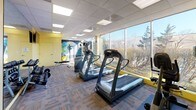 This fitness center is a thoughtfully designed, single-floor facility that offers a comprehensive workout environment for users of all fitness levels. The space features a modern drop-panel ceiling with recessed lighting, complemented by an abundance of natural light streaming through expansive floor-to-ceiling windows that frame views of the surrounding greenery and landscaping.

The interior is finished with a patterned, multi-tone carpet featuring bold orange accent lines, adding a dynamic visual energy to the space. The walls are painted in a warm yellow tone, accented by a vibrant motivational mural depicting an athletic silhouette, contributing to an energizing atmosphere.

The cardio zone is well-appointed with multiple Vision Fitness treadmills and an elliptical trainer, all positioned to take advantage of the outdoor views. A dual-pulley cable machine provides versatile strength training options, while a full rack of dumbbells in varying weights serves the free weights area. A weight bench is also available for supplementary exercises.

Full-length mirrors along one wall visually expand the space and assist users during workouts. Wall-mounted flat-screen televisions are positioned throughout the room for entertainment. Stability balls and additional accessories are stored near the mural wall, rounding out the facility's offerings. A private access door provides a dedicated entry point to the space.