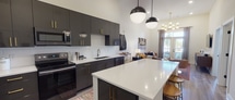Located at 3115 Shannon Rd in Durham, NC, this single-floor residence showcases a thoughtfully designed open-concept layout with soaring ceilings that amplify the sense of space throughout. The kitchen serves as a striking focal point, featuring sleek dark cabinetry accented with gold hardware, white quartz countertops, a subway tile backsplash, and stainless steel appliances including a French door refrigerator and range. A large central island with bar seating and globe pendant lighting bridges the kitchen and living areas seamlessly. The living room benefits from abundant natural light streaming through floor-to-ceiling glass doors that open to an outdoor balcony, complemented by a sophisticated brass multi-globe chandelier. Wood-look flooring flows throughout the main living spaces, adding warmth to the contemporary aesthetic. The primary bedroom offers a calm retreat with recessed lighting, a ceiling fan, and direct access to a well-appointed bathroom featuring a tiled walk-in shower, granite vanity, and an adjoining walk-in closet. A second bedroom provides a cozy, well-lit space with room for a workspace. The overall ambiance balances modern design with warm, livable comfort.