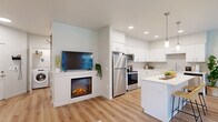 This well-appointed 2-bedroom, 2-bathroom residence combines modern design with functional living on a single floor. The open-concept layout seamlessly connects the living, dining, and kitchen areas, all unified by light hardwood-style flooring and a neutral color palette. The kitchen features white shaker cabinetry, quartz countertops, a subtle grey tile backsplash, and a full suite of stainless steel appliances, including a refrigerator, range, microwave, and dishwasher. A kitchen island provides additional prep and casual dining space, while pendant lighting adds a polished touch. The living area is highlighted by a built-in electric fireplace, recessed lighting, and large windows that invite natural light and views of the surrounding greenery. A dedicated dining area sits adjacent to the living space, also benefiting from window views. The primary bedroom offers a comfortable, carpeted retreat with a ceiling fan, a closet, and a window, while the second bedroom is equally well-sized. Both bathrooms are finished in clean, contemporary styling, with one featuring a glass-enclosed walk-in shower and wood-look flooring. A hallway connects the private and shared spaces efficiently, completing this cohesive and modern floor plan.