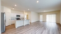 This single-story home is designed with a clean, modern aesthetic and a practical open-concept floor plan. The main living and kitchen area forms the heart of the home, where light wood-look plank flooring extends seamlessly throughout, reflecting recessed lighting and natural light from windows dressed with white blinds. The kitchen is well-appointed with crisp white cabinetry, granite countertops, and a central island that provides both workspace and casual seating. Stainless steel appliances, including a French door refrigerator, over-the-range microwave, and electric range, complete the kitchen. The adjacent living area is generously proportioned, offering an open, airy feel with neutral walls and a sliding glass door that allows additional natural light. A dedicated laundry room includes built-in shelving and connections for a washer and dryer. The primary bathroom features an extended double vanity with white cabinetry and a walk-in glass shower, while a secondary bathroom offers a full tub-shower combo. A spacious walk-in closet with wrap-around shelving and carpeted flooring provides ample storage. Throughout, the home maintains a consistent, neutral palette that creates a bright and cohesive atmosphere.