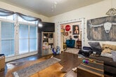 This single-floor home offers a warm and eclectic living experience across several distinct spaces. The main living area is a generous room featuring rich hardwood floors, a ceiling fan, and large windows dressed with draped curtains that allow natural light to filter through horizontal blinds. A wall-mounted flat-screen television, open storage shelving, and a cowhide-style area rug add to the room's distinct personality. French doors open into an adjoining sitting room or secondary lounge, decorated with wall art and string lights, creating a cozy, personalized nook with additional seating. The two bedrooms are comfortably sized, each outfitted with ceiling fans, multiple windows with layered curtain treatments, and hardwood flooring. Bohemian-inspired wall décor, including a macramé dreamcatcher and metal floral accents, gives the bedrooms a relaxed, creative character. The kitchen follows a compact galley configuration with white cabinetry, open wall shelving for dishware, dark countertops, and a practical hanging rack for cookware. A gas range and sink are efficiently positioned along the narrow layout. Throughout the home, the interplay of hardwood floors, natural light from multiple windows, and eclectic décor creates a lived-in, expressive atmosphere full of individual character.
