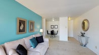 This single-floor residence offers a thoughtfully arranged open-concept layout that seamlessly connects the living and dining areas, creating a cohesive and functional living environment. The living area is anchored by a bold teal accent wall that adds a vibrant pop of color against the neutral white ceiling and walls, lending the space a modern yet inviting character. Plush wall-to-wall carpeting runs throughout, adding warmth and comfort underfoot. Natural light complements the flush ceiling fixtures to keep the space feeling bright and airy. Framed wall art enhances the décor, contributing to a curated, finished aesthetic. The dining area is situated just beyond the living space, defined by a sleek glass-top dining table surrounded by contemporary black chairs, creating a clean and stylish setting for meals. The transition between the two areas flows naturally, making the overall space feel open and connected. A private entry door is visible in the background, offering a sense of separation from the outside. The overall ambiance is tidy and modern, with a cohesive color palette of teal, white, and neutral tones that gives the home a fresh and contemporary feel throughout.