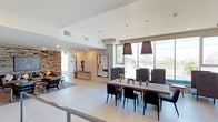 This single-floor space showcases a thoughtfully designed open-plan layout that seamlessly connects a dining area, lounge, and additional amenity spaces. The dining area is anchored by a solid wood rectangular table surrounded by black upholstered chairs, illuminated by a pair of striking oversized black pendant lights. Large floor-to-ceiling windows flood the space with natural light and provide open exterior views, enhancing the airy, contemporary atmosphere.

A standout feature throughout is the reclaimed wood stone-textured accent wall, which adds warmth and visual depth to the otherwise clean, minimalist palette of white walls and polished concrete-style flooring. The lounge area is furnished with a generously sized gray sectional sofa, black leather accent chairs, and a metal-framed coffee table, centered around a wall-mounted flat-screen television. Curated artwork, including bold abstract canvases and sculptural pieces, is displayed throughout, lending the space a gallery-like quality.

Adjacent to the dining area, a kitchenette or refreshment station is visible behind glass partitioning, adding functional versatility. The space is ADA accessible, and recessed lighting complements the natural light throughout, maintaining a bright and welcoming ambiance.