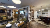 This single-floor space showcases a thoughtfully designed open-concept layout that seamlessly blends modern aesthetics with warm, inviting elements. At the heart of the main lounge area, a striking feature wall clad in dark textured tile serves as a dramatic focal point, housing a wall-mounted television above a sleek linear fireplace. Flanking this centerpiece, open white shelving units with dark wood accents provide both visual interest and functional display space, effectively defining zones within the open floor plan.

Rich, wide-plank hardwood flooring runs throughout, grounding the space with warmth and continuity. Exposed brick accent walls add an industrial character, complementing the contemporary furnishings, including plush olive-green seating and a minimalist console table styled with decorative objects. Recessed lighting and oversized circular pendant fixtures illuminate the space, while natural light filters in from windows visible toward the rear.

Beyond the main seating area, a billiards table nestled beneath ambient lighting suggests a dedicated recreation zone. A separate, partially enclosed room is visible to the right, hinting at additional functional space. The overall atmosphere is sophisticated yet approachable.