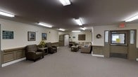 Located at 1000 Nicholas Brooks Ct in New Windsor, New York, this single-floor facility offers a thoughtfully arranged and welcoming interior designed for comfort and community. The main lounge area is generously proportioned, featuring plush upholstered seating arrangements including armchairs and a sofa grouped around a central coffee table, creating an inviting space for gathering and conversation. The room is finished with neutral-toned carpet flooring and cream-colored walls adorned with framed artwork, a decorative wreath, and a textured wall sculpture, adding warmth and character to the environment. Overhead, flush-mounted fluorescent lighting provides consistent, even illumination throughout the space. A dark ceiling treatment creates a subtle contrast against the lighter walls and furnishings, lending the room a grounded, cozy ambiance. Fresh floral arrangements add a welcoming touch to the seating areas. Toward the rear of the space, a doorway leads to additional rooms, suggesting further functional areas beyond the main lounge. The overall design reflects a practical yet comfortable aesthetic, well-suited for a community or care-oriented facility seeking to provide residents and visitors with a calm and accommodating environment.